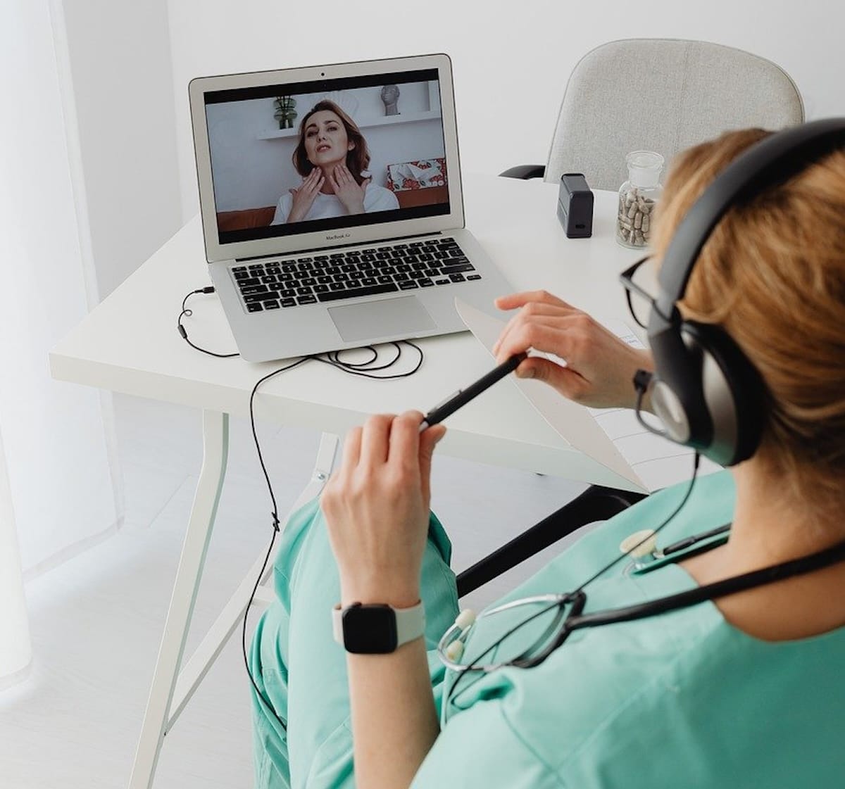 Medical professional in medical scrubs wearing headphones and stethoscope watching telehealth session via laptop computer