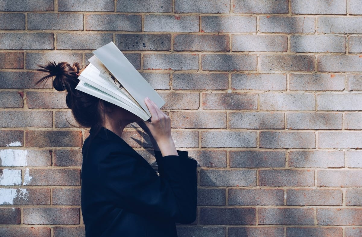 Individual student standing beside a brick wall with a textbook covering their face outdoors