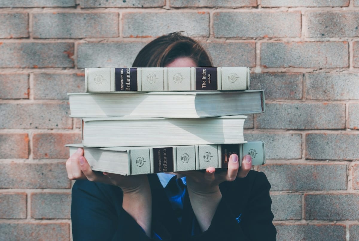 Individual outdoors against a brick wall holding a stack of four books