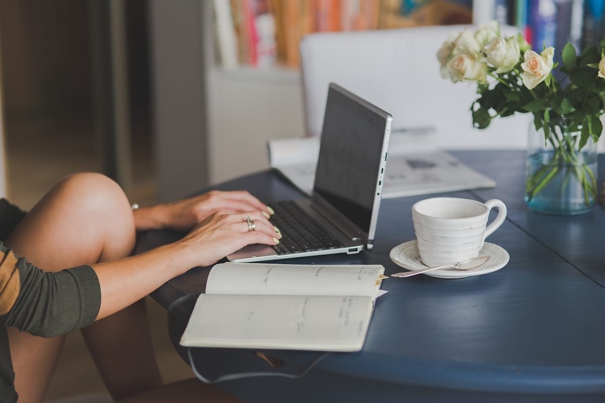 Individual sitting at a table with a laptop computer with notebooks and cup